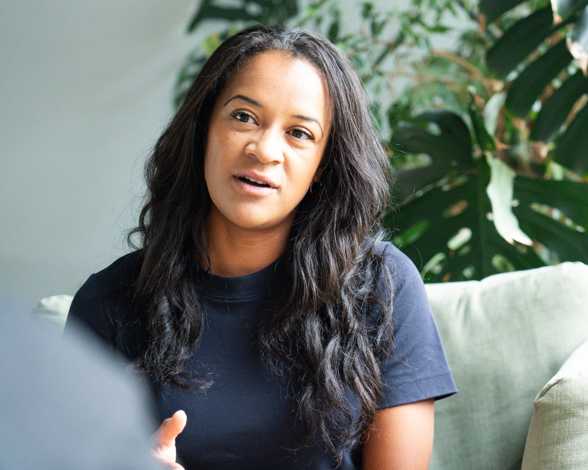 A woman with long dark hair sits on a couch, talking about feedback for career growth, with green plants in the background.