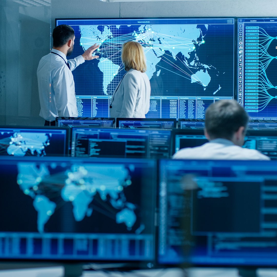 Two people in a control room analyze a world map with data lines on a large SCC Core screen, surrounded by monitors displaying digital information.