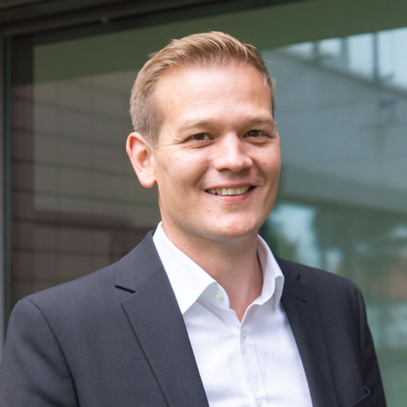 A man in a dark suit and white shirt smiles while standing outside near a glass window, perhaps on his way to an interview for Full Stack Developer jobs.