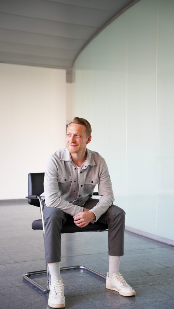 A man with light hair sits on a black chair in a modern indoor space, dressed smartly as a Consultant. Wearing a light gray shirt, dark pants, and white shoes, he smiles confidently—ready to discuss Supply Chain Digitalisierung.