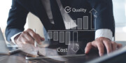 A person in business attire examines documents at a desk; overlaid graphics show quality increasing and cost decreasing with bar charts and percentage symbols.