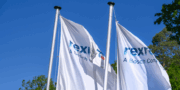 Three white flags with the rexroth A Bosch Company logo are flying against a clear blue sky, with some green trees visible in the background.