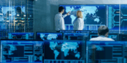 Two people analyze data on a large world map display in a control room with multiple computer monitors and digital information screens.