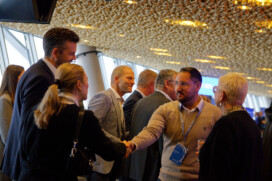 People in business attire shake hands and converse at a professional networking event in a modern indoor venue.