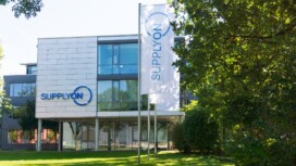 Modern office building with SupplyOn signage and logo on the facade and flag, surrounded by trees and green lawn.