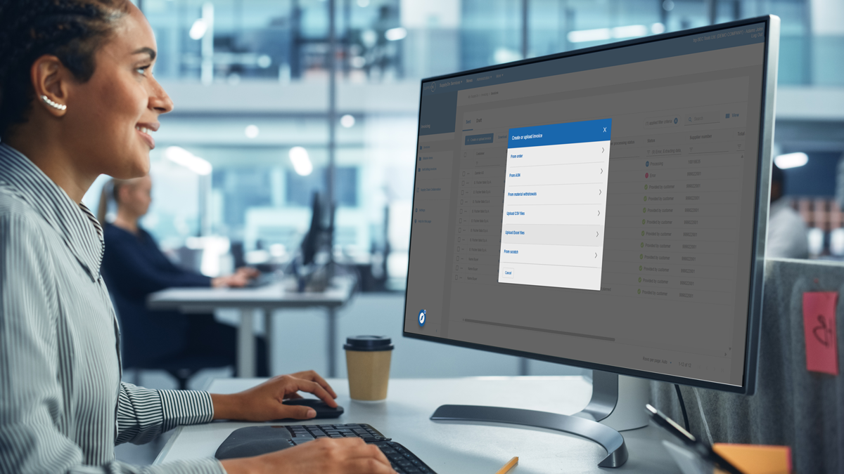 A person at a desk uses a desktop computer with a large monitor displaying a drop-down menu; office setting visible in the background.