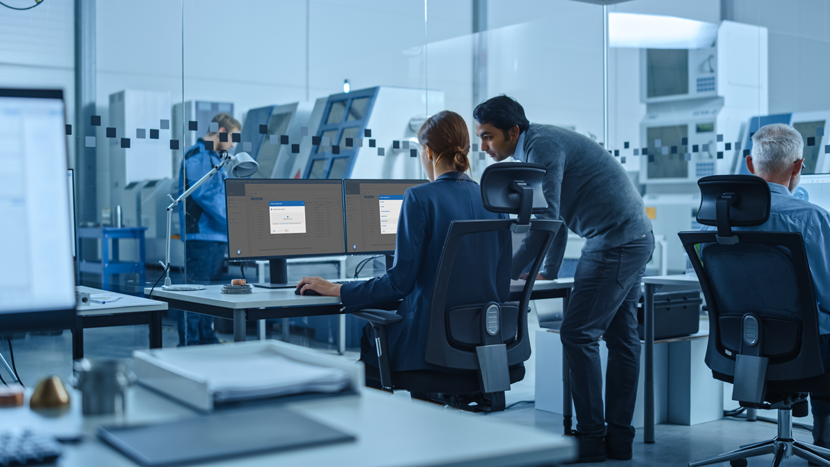 Four people work in a modern office with large computer monitors; two collaborate at a desk whilst others focus on their screens in the background.
