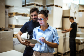 Two people review information on a tablet in a warehouse, surrounded by stacked pallets and boxes, whilst another person works in the background.
