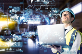 Man in safety vest and hard hat holding a laptop, looking at floating digital data graphics representing global technology and connectivity in an industrial setting.