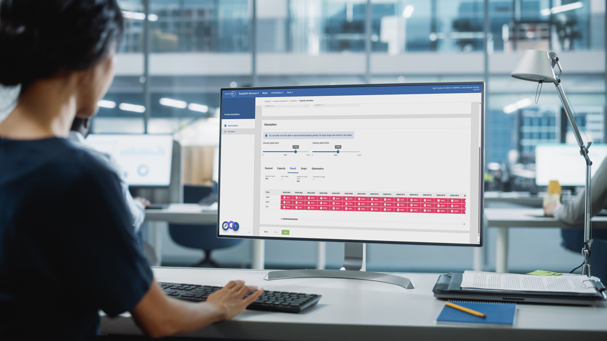 Person working at a desk in a modern office, viewing a scheduling or planning interface with a calendar grid displayed on a large computer monitor.