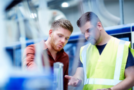 Two men in a factory setting discuss and examine machinery; one wears a brown shirt, the other a high-visibility vest.