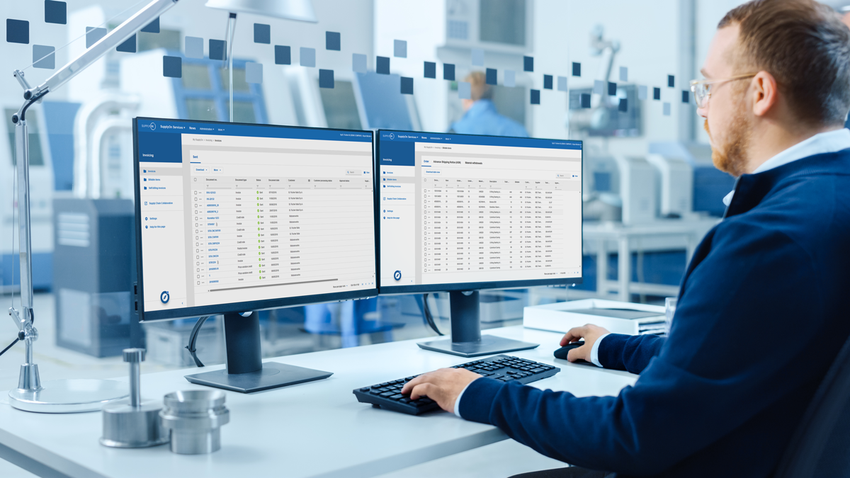 A person sits at a desk in an office using dual monitors displaying data tables and software interfaces, with industrial equipment visible in the background.