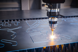 A laser cutting machine precisely cuts shapes into a metal sheet, with sparks visible at the cutting point.