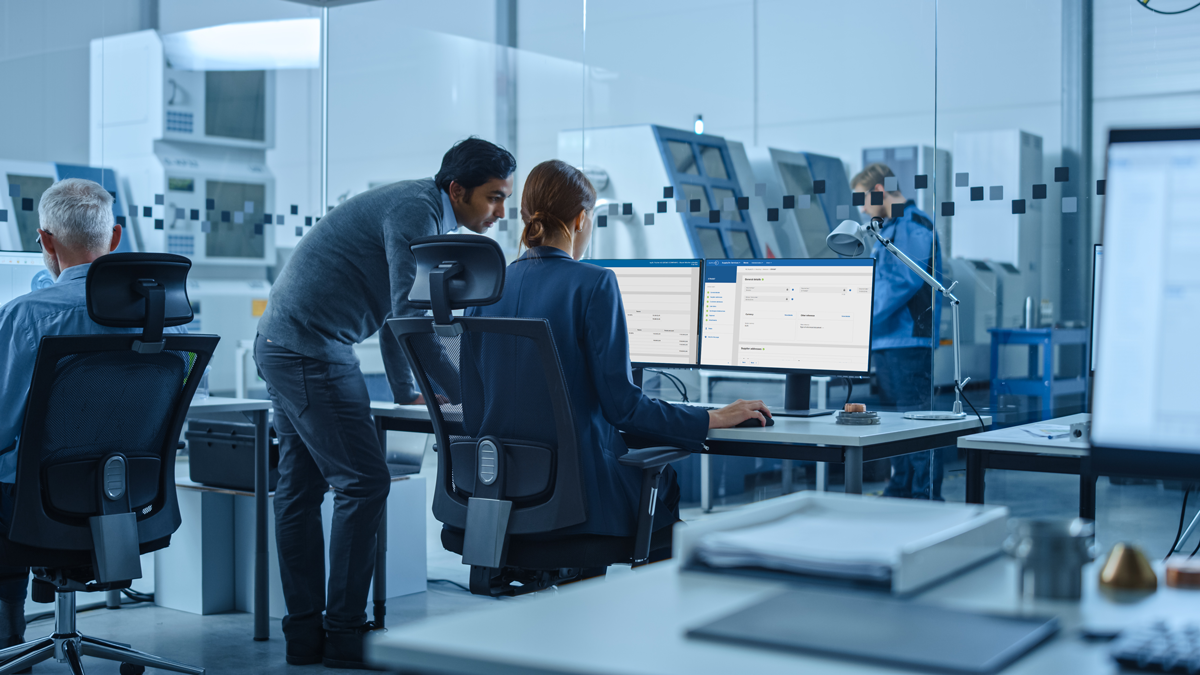 Three people work at computer desks in a modern office with glass walls; another person stands in the background. Monitors display data, and office equipment is visible.