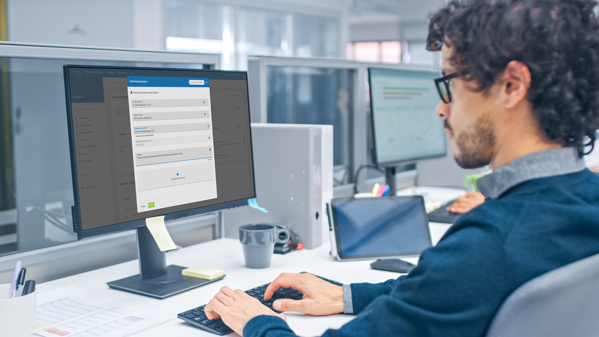 A person with curly hair and glasses works at a desktop computer in a modern office, filling in an online form on the screen.