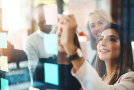 A group of people stands by a glass wall with colourful sticky notes as a woman in the foreground writes on one of the notes with a marker.