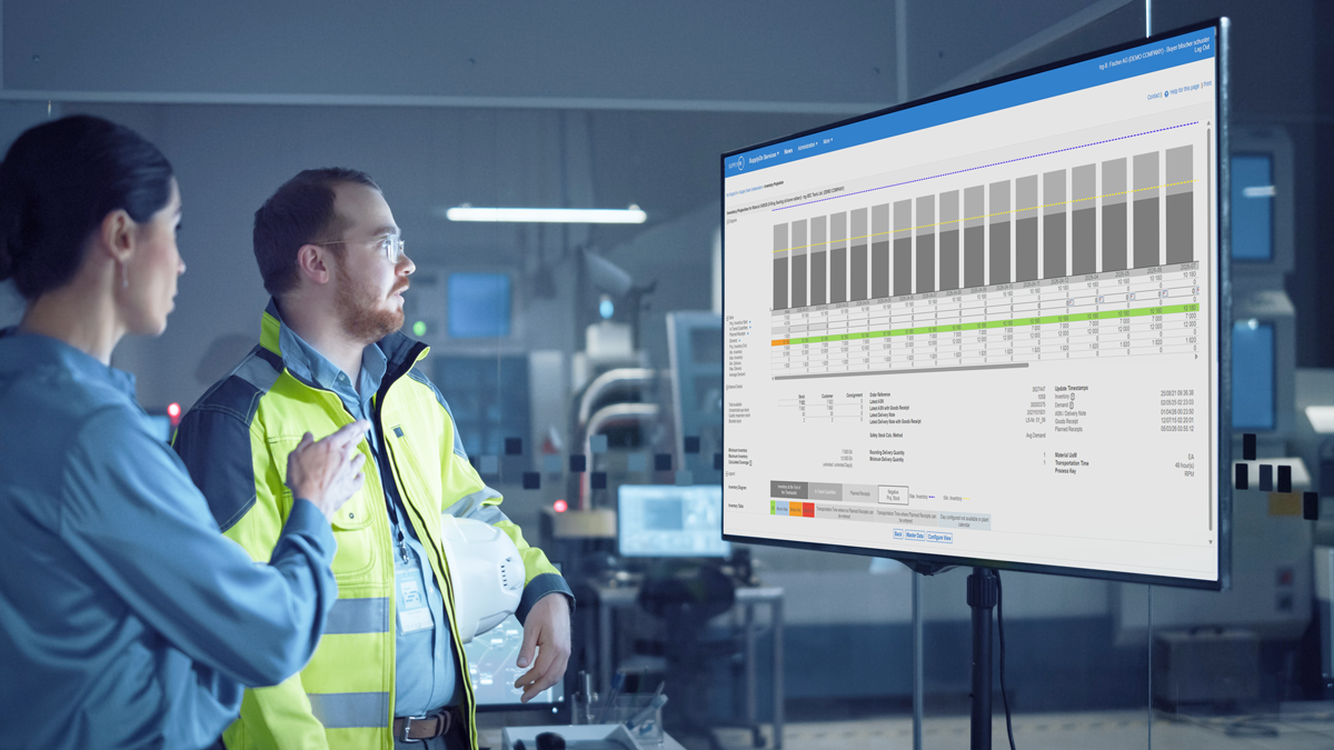 Two people in work attire review a large screen displaying a detailed data chart with graphs and tables in a modern industrial setting.