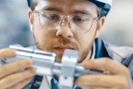 A person wearing spectacles and a hard hat closely examines a metal mechanical part held in their hands.