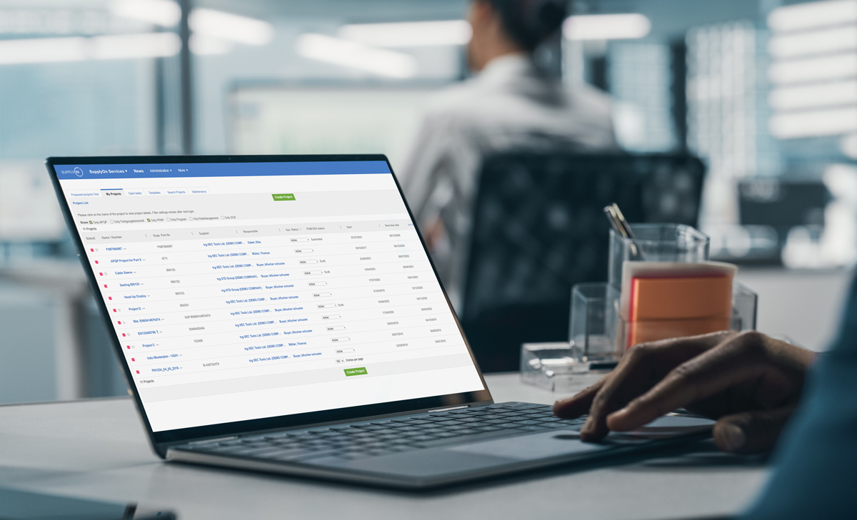 Person working on a laptop displaying a webpage with a table of shipment records in an office setting; blurred figure and desk items visible in the background.
