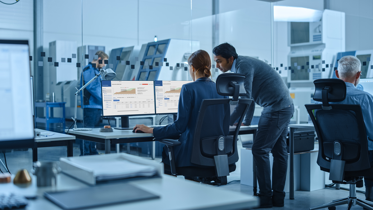 Office workers at computer desks analyse data charts on monitors in a modern workspace, while others collaborate and operate machinery in the background.