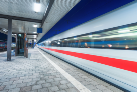 A high-speed train with a red stripe moves past an empty station platform at night, creating a motion blur effect.