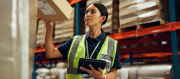 A woman wearing a high-visibility vest holds a parcel and uses a tablet whilst working in a warehouse with shelves and boxes in the background.