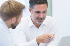 Two men in white shirts are sitting together, with one pointing at a laptop screen whilst they have a discussion.