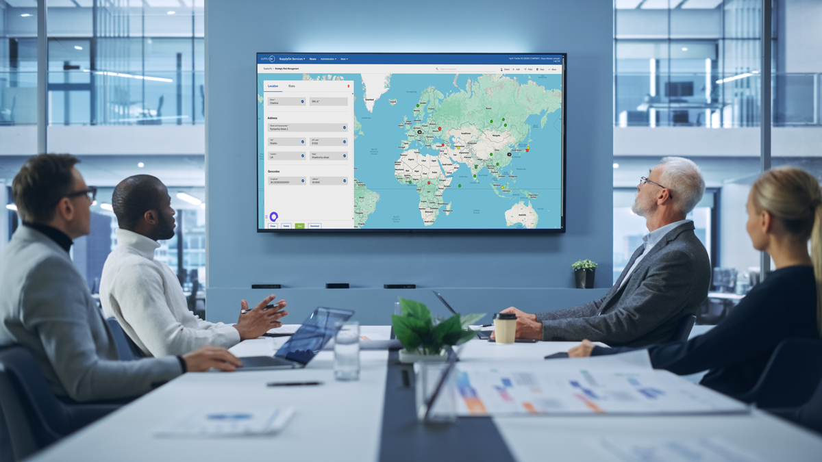 Four people sit at a conference table in an office, viewing a world map with data displays on a large wall-mounted screen.