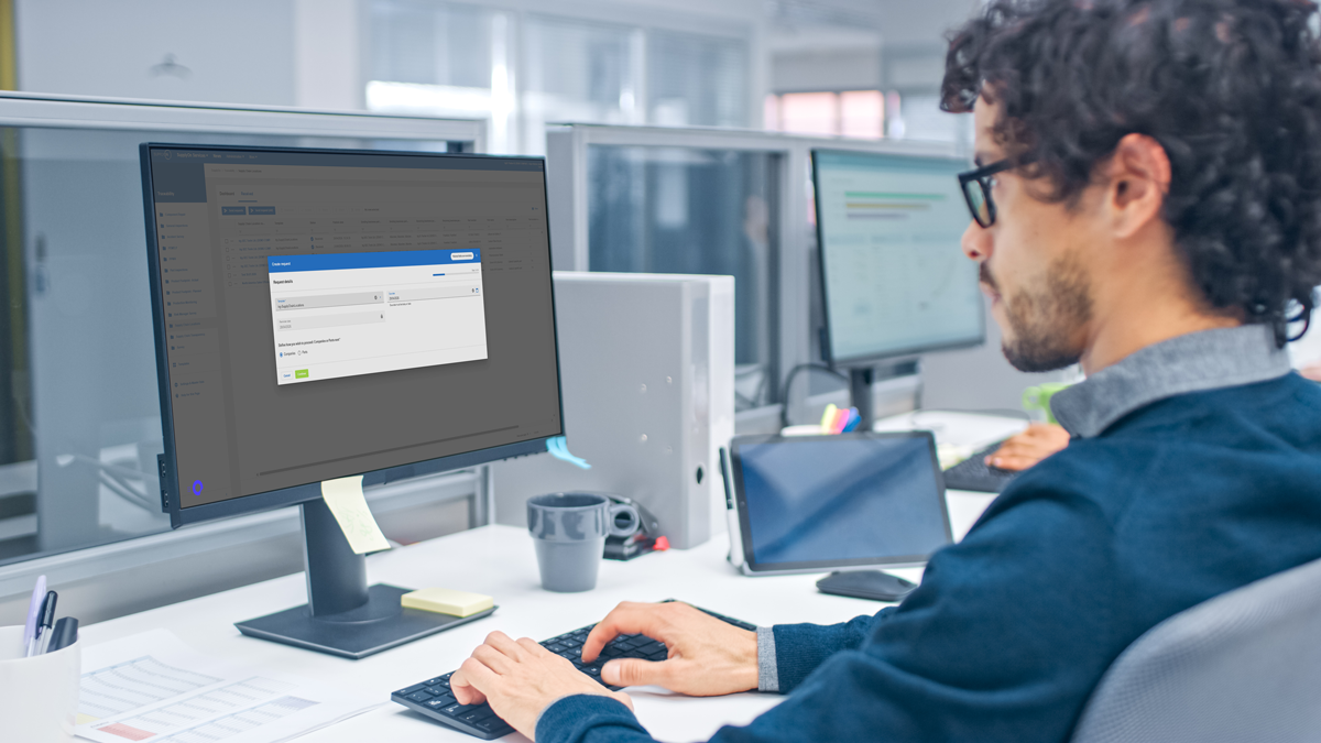 A man sits at a desk in a modern office, typing on a keyboard and viewing a form on a large desktop monitor.