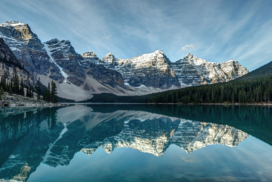Snow-capped mountains and evergreen trees are reflected in a calm, turquoise lake under a partly cloudy sky.