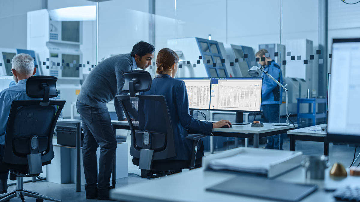 Office workers sitting at desks with computer monitors displaying data, while a man stands and talks to a seated woman; machinery and another person are visible in the background.