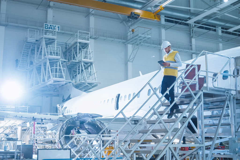 An engineer wearing a hard hat and yellow vest walks down stairs beside a commercial aeroplane inside an aircraft maintenance hangar.