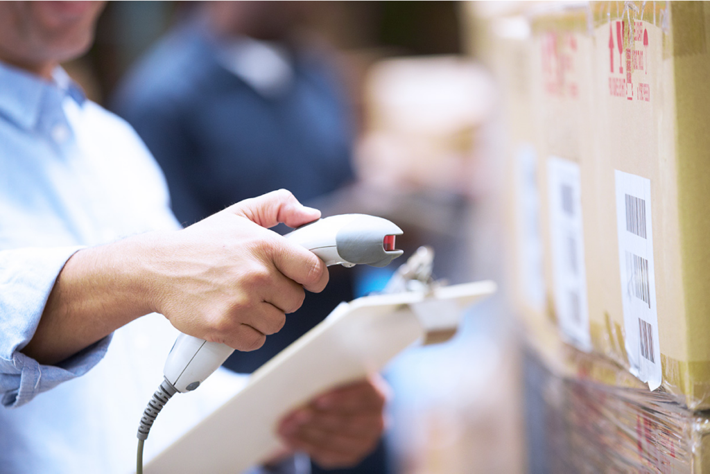 Person holding a clipboard uses a barcode scanner to scan labels on stacked cardboard boxes in a warehouse.