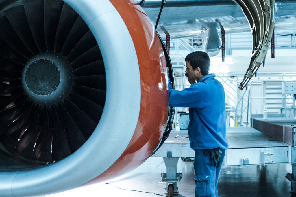 A technician in blue workwear inspects or repairs a jet engine in a brightly lit aircraft maintenance hangar.