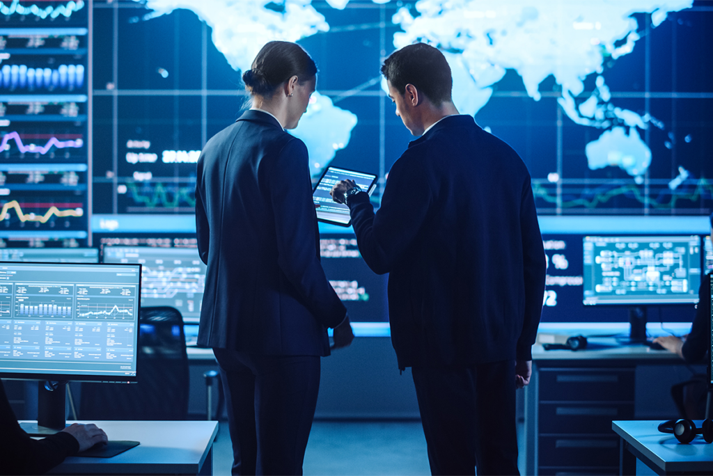 Two people in business attire review data on a tablet in a control room with large digital screens displaying world maps and charts.