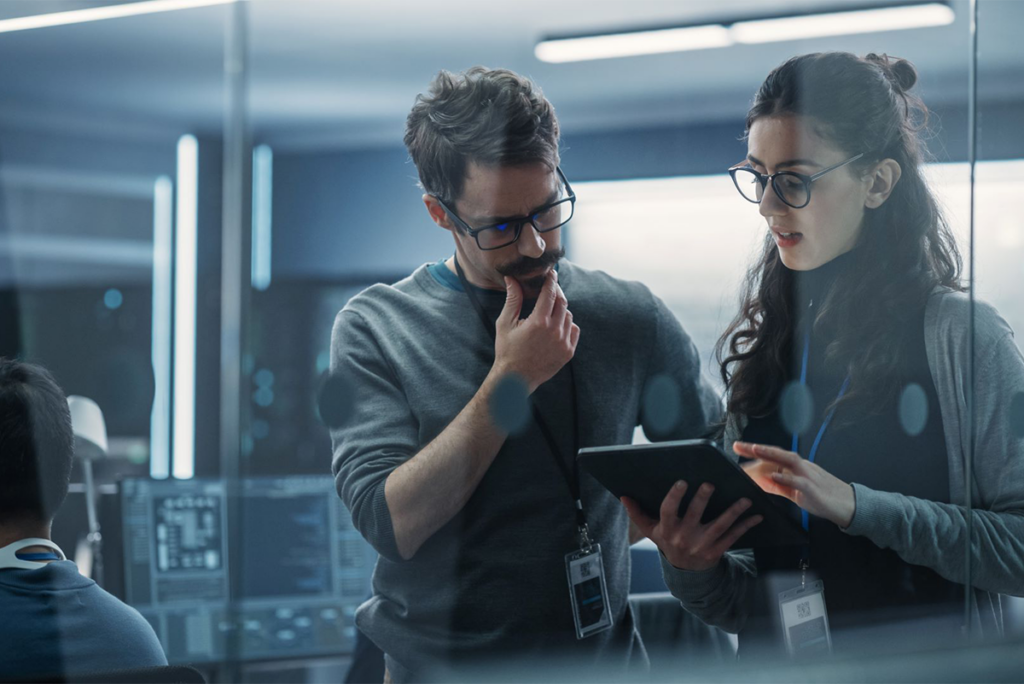Two people wearing glasses and ID badges stand in an office, reviewing information on a tablet together, with computer monitors visible in the background.