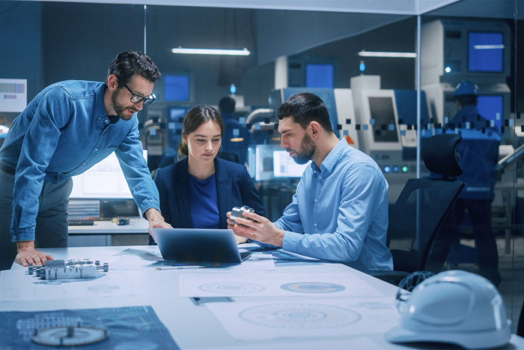Three professionals in an office review a 3D-printed part and data on a laptop, surrounded by technical documents and engineering equipment.