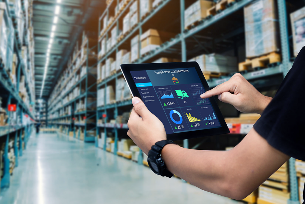Person holding a tablet displaying warehouse management data and analytics in a large warehouse with shelves of boxes and goods.