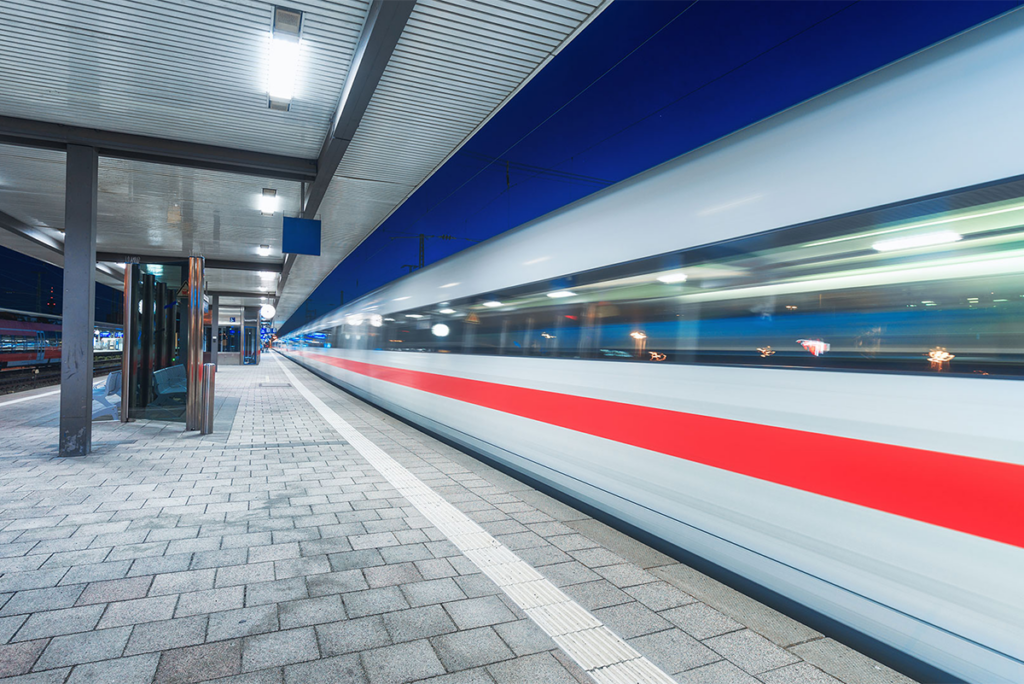 A high-speed train with a red stripe moves rapidly through an empty, modern railway station at dusk.