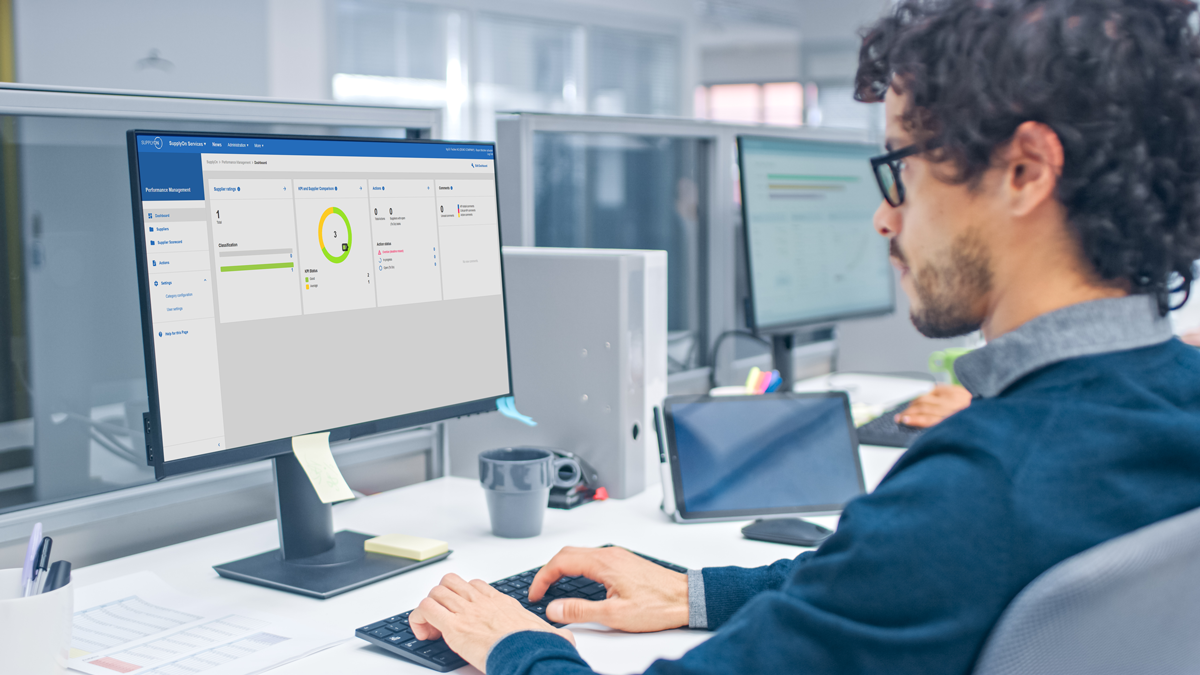 A person with curly hair and glasses works at a desk, looking at a computer monitor displaying a dashboard with graphs and charts in an office setting.