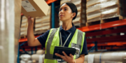 A woman in a high-visibility vest holds a parcel and checks information on a tablet in a warehouse.