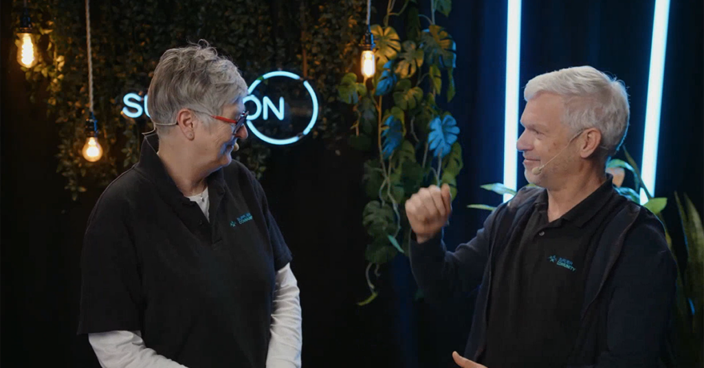 Two people wearing microphones and black shirts communicate using sign language in front of plants and neon lights.