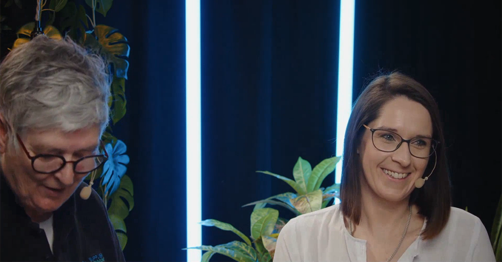 Two people wearing microphones sit indoors, with plants and vertical blue lights in the background. One person is smiling; the other is looking down.