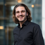 A young man with shoulder-length brown hair and glasses, wearing a black shirt, smiles whilst standing outdoors in front of a glass building.