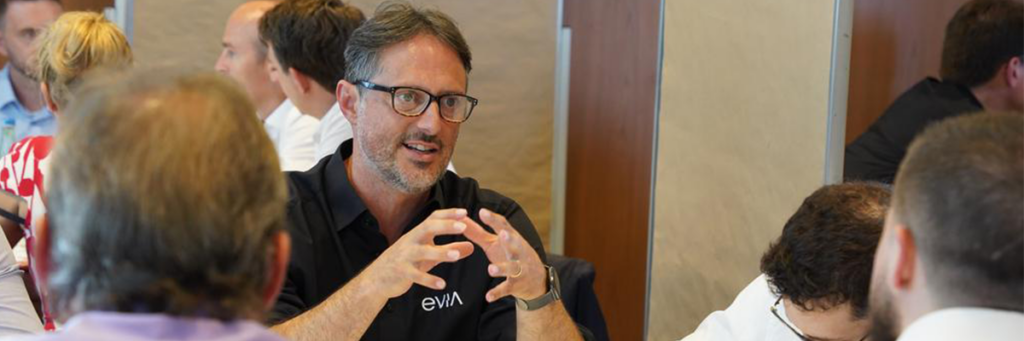 A man in glasses speaks and gestures with his hands whilst seated at a table with several others in a meeting focused on Defence Readiness and discussion.