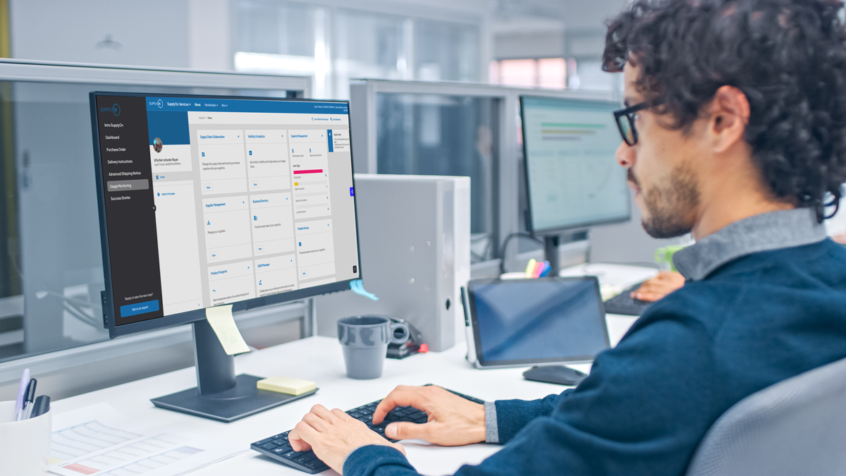 A person with curly hair and glasses works at a desk, viewing a digital dashboard on a large computer monitor in a modern office setting.